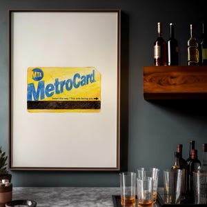 May include: A framed artwork featuring a yellow MTA MetroCard with blue lettering and the text "Insert this way / This side facing you." The artwork is displayed on a white background, with a dark frame. The image is set against a dark gray wall.