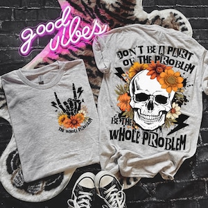 以下が含まれることがあります： 骸骨と花のデザインが施されたグレーのTシャツ2枚。シャツには「問題の一部になるな、問題全体になれ」という文字が書かれている。