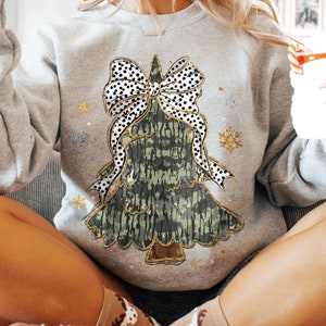 Può includere: Una felpa grigia con un design di albero di Natale verde e oro. L'albero ha un grande fiocco a pois neri e bianchi in cima. La felpa è decorata con glitter dorati e fiocchi di neve.