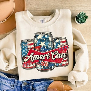 May include: Cream-colored sweatshirt featuring a graphic of three beverage cans with a patriotic design of stars and stripes. The cans are arranged with a red banner that reads "Ameri Can" in a distressed font. The sweatshirt is folded on a wooden surface.