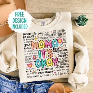 May include: A beige sweatshirt with a colorful graphic print that says "Mama It's Okay". The graphic includes phrases like "To not be okay", "To say no", "To enjoy the silence", "To feel overwhelmed", "To put yourself first", "To seek joy", "To set boundaries", "To enjoy hobbies", "To make mistakes", "To embrace imperfection", "To prioritize your health", "To change your mind", "To ask for help", and "To not always be strong".