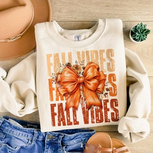 Puede incluir: Una sudadera de color crema con el texto "Fall Vibes" en naranja, marrón y amarillo. El texto está superpuesto y tiene un aspecto desgastado. Un gran lazo naranja con un diseño floral está centrado en la sudadera.