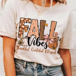 Pode incluir: Uma camiseta bege com um design gráfico que diz "Fall Vibes" em letras marrons, laranja e brancas com um padrão de outono. O texto "Thankful Grateful Blessed" está escrito em marrom abaixo do gráfico.
