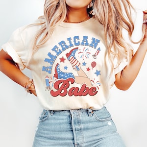 May include: A white t-shirt with a graphic of a woman's legs wearing cowboy boots with an American flag design. The text "American Babe" is printed in red and white on the shirt.