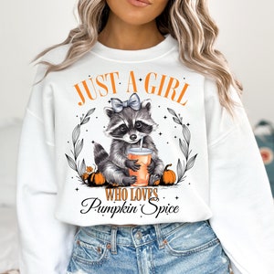 May include: White sweatshirt with a graphic of a raccoon wearing a bow and holding a pumpkin spice drink. The text "Just a girl who loves pumpkin spice" is printed below the raccoon.
