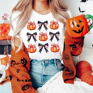 Puede incluir: Camiseta blanca con un patr&oacute;n de calabazas naranjas con lazos negros y rosas. Las calabazas est&aacute;n dispuestas en una cuadr&iacute;cula.