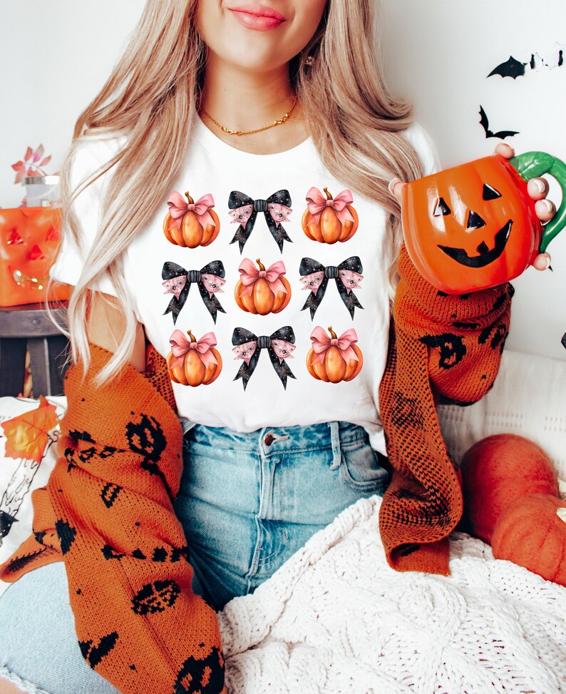Puede incluir: Camiseta blanca con un patr&oacute;n de calabazas naranjas con lazos negros y rosas. Las calabazas est&aacute;n dispuestas en una cuadr&iacute;cula.