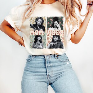 May include: A white t-shirt with a vintage-style graphic featuring four women's faces in squares with floral backgrounds. The text "MOM VIBES" is printed in pink letters below the images.