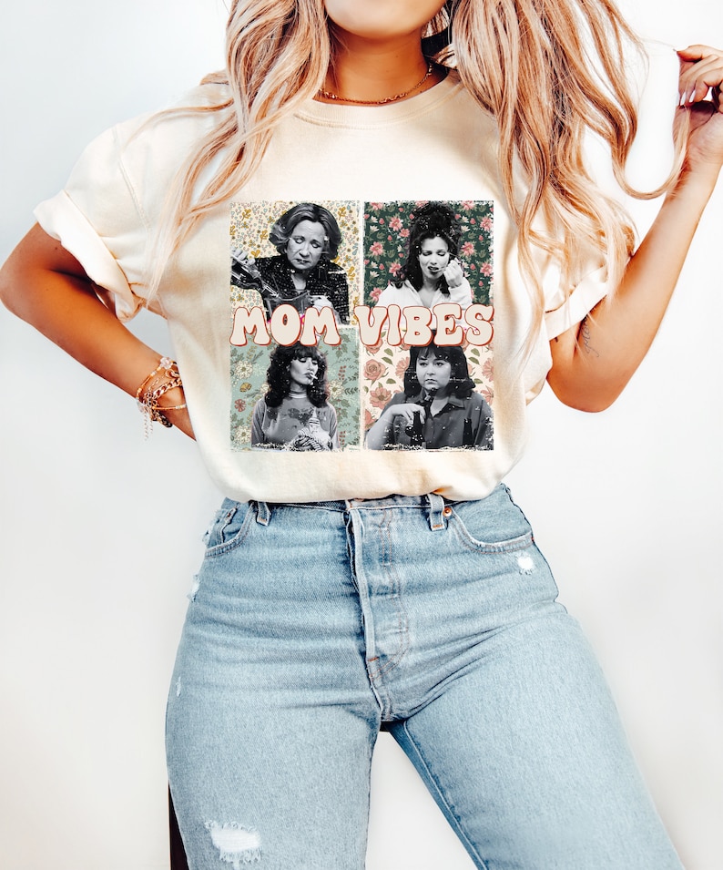 May include: A white t-shirt with a vintage-style graphic featuring four women's faces in squares with floral backgrounds. The text "MOM VIBES" is printed in pink letters below the images.