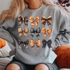 May include: A grey sweatshirt featuring a pattern of orange and black bows. The bows are in various styles, including polka dots, plaid, and leopard print.