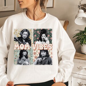 May include: White sweatshirt with a graphic print of four women's faces with floral backgrounds. The text "MOM VIBES" is printed in a retro font.