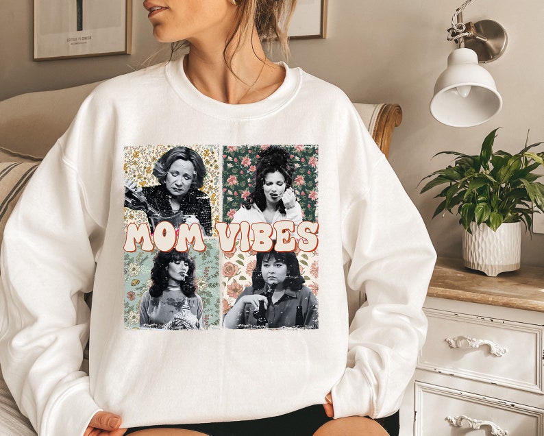 May include: White sweatshirt with a graphic print of four women's faces with floral backgrounds. The text "MOM VIBES" is printed in a retro font.