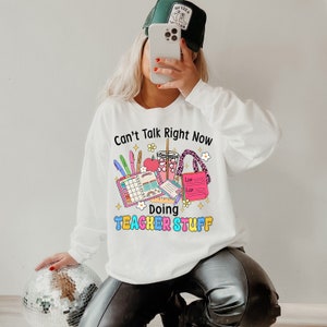May include: White long-sleeved sweatshirt featuring a colourful graphic. The graphic reads "Can't Talk Right Now Doing Teacher Stuff" with illustrations of pens, a planner, a drink, and a list. The sweatshirt is a casual style.