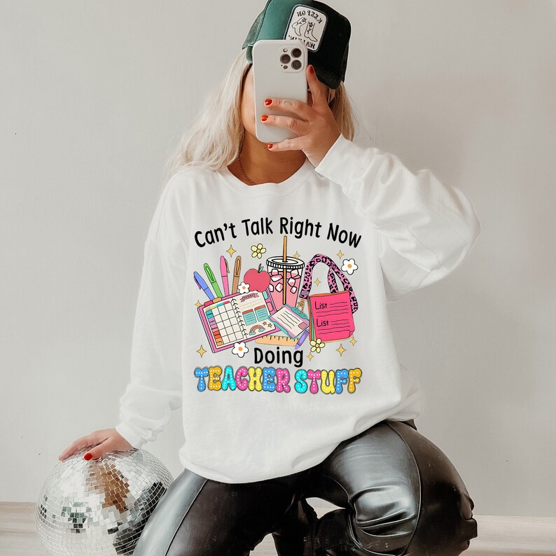 May include: White long-sleeved sweatshirt featuring a colourful graphic. The graphic reads "Can't Talk Right Now Doing Teacher Stuff" with illustrations of pens, a planner, a drink, and a list. The sweatshirt is a casual style.