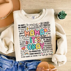 May include: A cream-colored sweatshirt with a colorful graphic print that says "To not be okay, to say no, to feel overwhelmed, to enjoy the silence, to put yourself first, Mama it's okay, to set boundaries, to enjoy hobbies, to make mistakes, to embrace imperfection, to prioritize your health, to change your mind, to ask for help, to not always be strong".