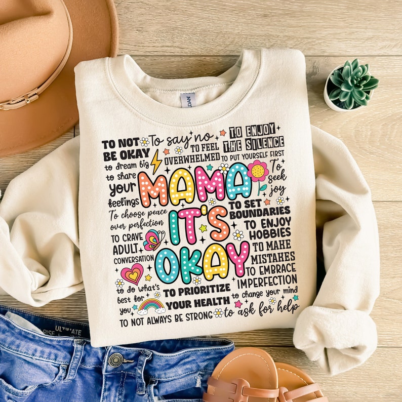 May include: A cream-colored sweatshirt with a colorful graphic print that says "To not be okay, to say no, to feel overwhelmed, to enjoy the silence, to put yourself first, Mama it's okay, to set boundaries, to enjoy hobbies, to make mistakes, to embrace imperfection, to prioritize your health, to change your mind, to ask for help, to not always be strong".