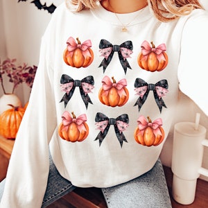 Puede incluir: Sudadera blanca con un patr&oacute;n de calabazas naranjas con lazos rosas y lazos negros con lunares blancos y rosas.
