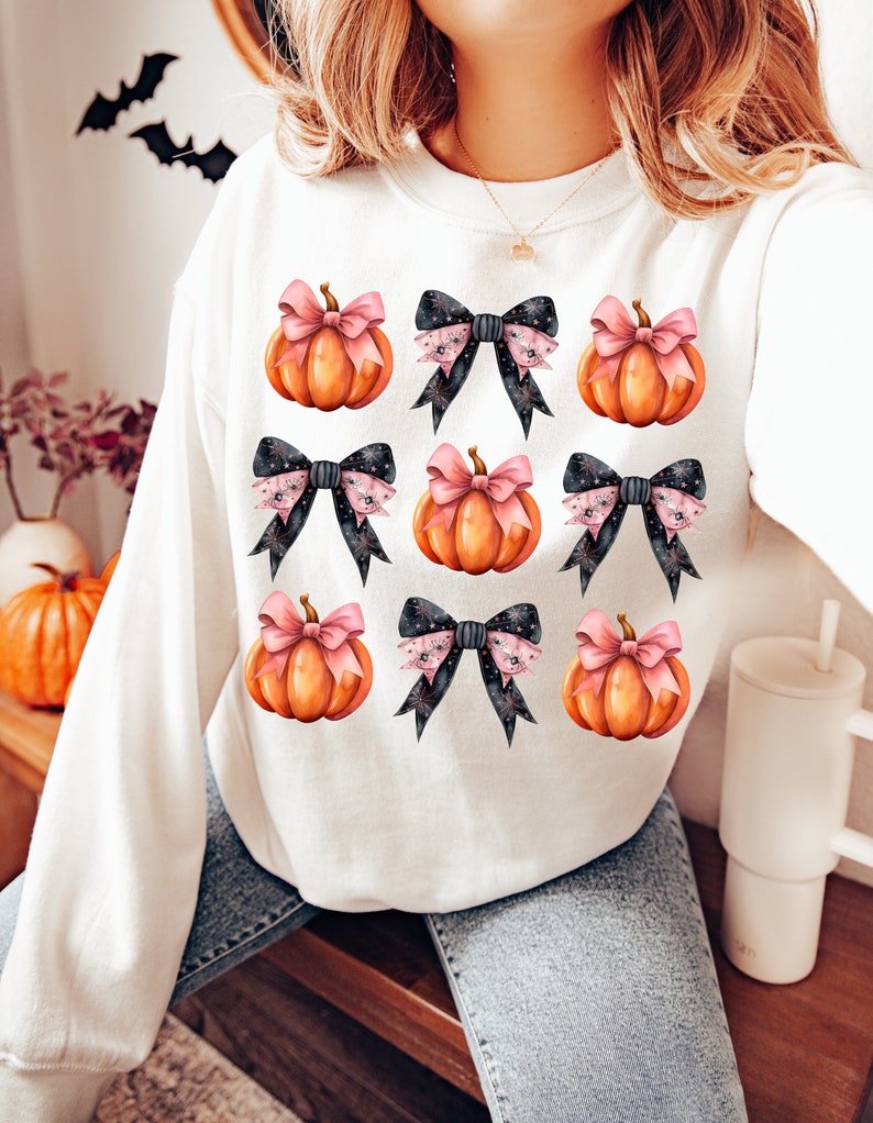 Puede incluir: Sudadera blanca con un patr&oacute;n de calabazas naranjas con lazos rosas y lazos negros con lunares blancos y rosas.