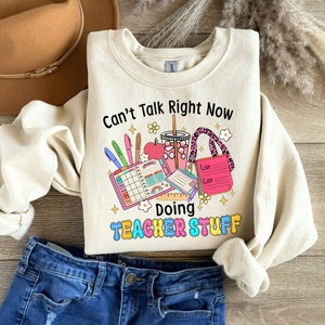 May include: Cream-coloured sweatshirt featuring the phrase "Can't Talk Right Now Doing Teacher Stuff". The design includes a planner, pens, an apple, a drink, and a tote bag. The sweatshirt is shown with blue jeans and a brown hat.