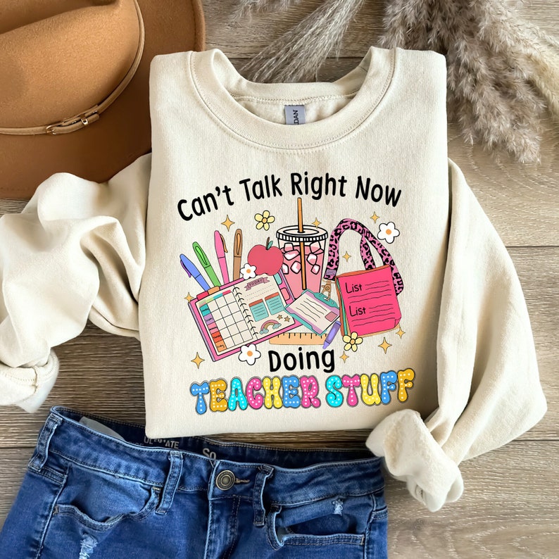 May include: Cream-coloured sweatshirt featuring the phrase "Can't Talk Right Now Doing Teacher Stuff". The design includes a planner, pens, an apple, a drink, and a tote bag. The sweatshirt is shown with blue jeans and a brown hat.