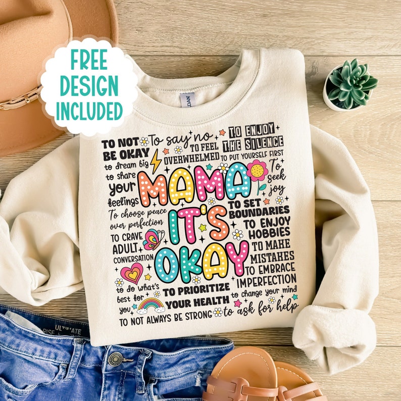 May include: A beige sweatshirt with a colorful graphic print that says "Mama It's Okay". The graphic includes phrases like "To not be okay", "To say no", "To enjoy the silence", "To feel overwhelmed", "To put yourself first", "To seek joy", "To set boundaries", "To enjoy hobbies", "To make mistakes", "To embrace imperfection", "To prioritize your health", "To change your mind", "To ask for help", and "To not always be strong".
