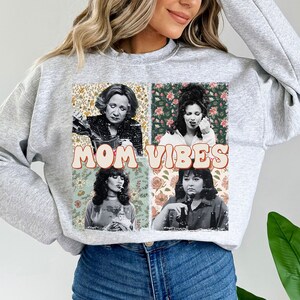 May include: Gray sweatshirt with a vintage-style graphic featuring four women's faces and the text "MOM VIBES" in colorful letters. The graphic is surrounded by floral patterns.