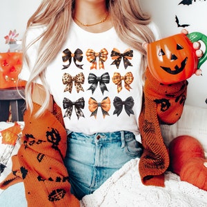 May include: A white t-shirt featuring a design of 12 black, orange and gold bows. The bows are arranged in four rows of three bows each.