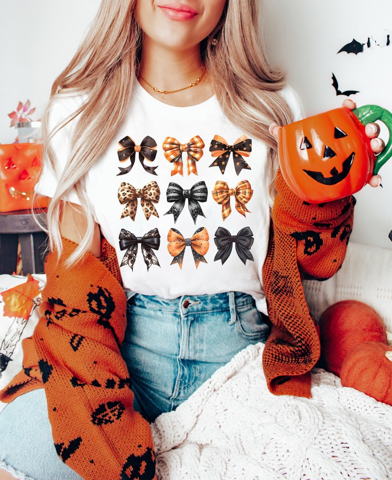 May include: A white t-shirt featuring a design of 12 black, orange and gold bows. The bows are arranged in four rows of three bows each.