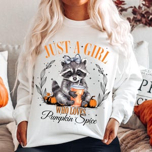 May include: White sweatshirt with a raccoon graphic holding a cup of coffee and the text "Just a girl who loves pumpkin spice".