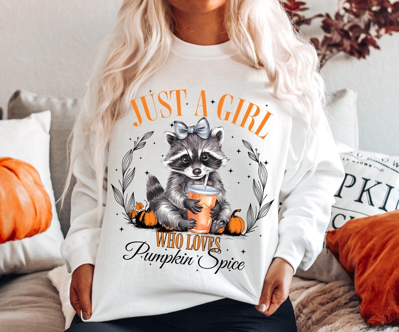 May include: White sweatshirt with a raccoon graphic holding a cup of coffee and the text "Just a girl who loves pumpkin spice".