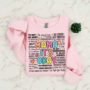 May include: Pink sweatshirt with a colorful graphic print that says "Mama It's Okay" and includes phrases like "To not be okay", "To say no", "To enjoy the silence", "To feel overwhelmed", "To put yourself first", "To seek joy", "To set boundaries", "To enjoy hobbies", "To make mistakes", "To embrace imperfection", "To prioritize your health", "To change your mind", "To ask for help".