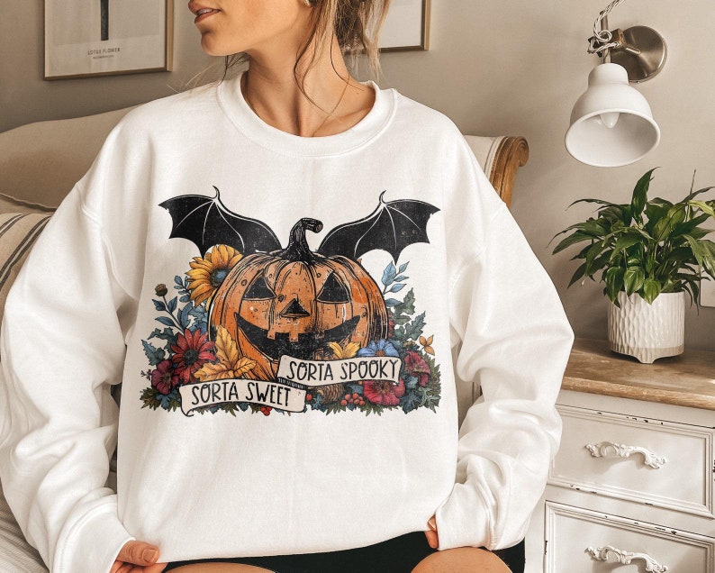 May include: White sweatshirt with a graphic of a jack-o'-lantern with bat wings and flowers. The text "Sorta Sweet" and "Sorta Spooky" is printed on a banner below the pumpkin.