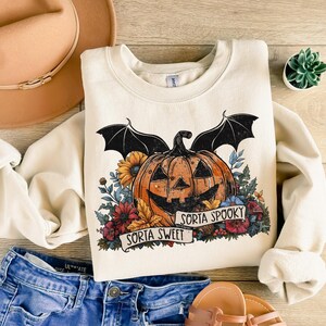 May include: A cream-colored sweatshirt with a graphic of a jack-o'-lantern with bat wings and flowers. The text "Sorta Sweet" and "Sorta Spooky" is printed on a banner below the jack-o'-lantern.