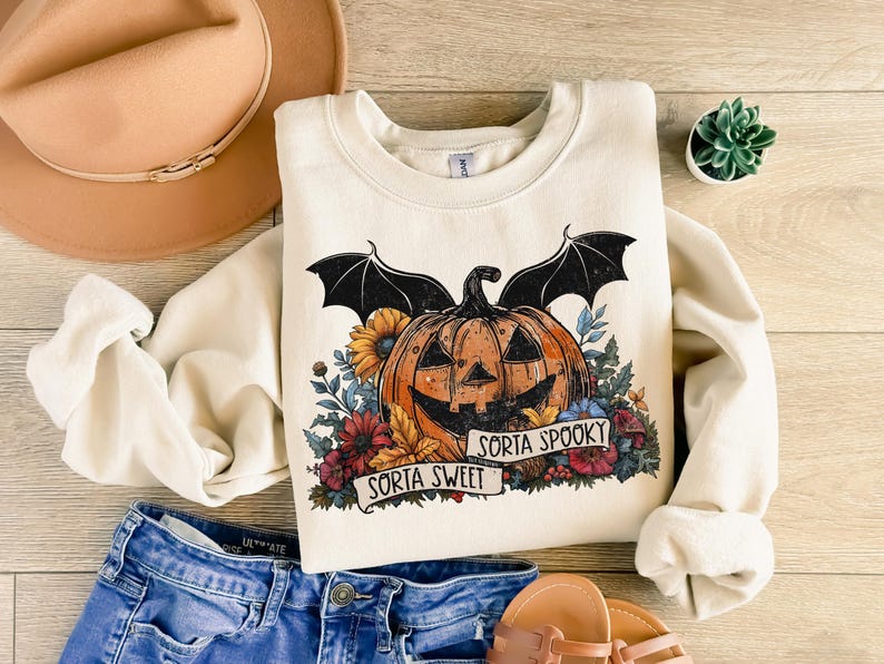 May include: A cream-colored sweatshirt with a graphic of a jack-o'-lantern with bat wings and flowers. The text "Sorta Sweet" and "Sorta Spooky" is printed on a banner below the jack-o'-lantern.