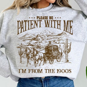 May include: Gray sweatshirt with a brown graphic print of a stagecoach pulled by horses. The text on the sweatshirt reads "Please be patient with me I'm from the 1900s".