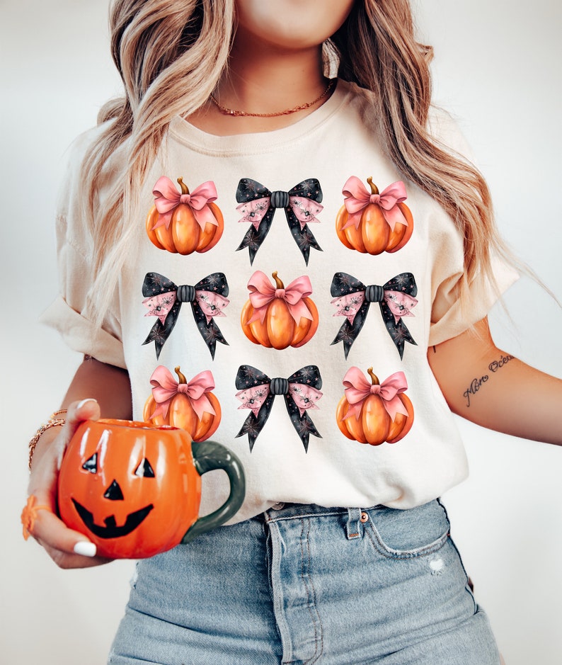 Puede incluir: Una camiseta blanca con un patr&oacute;n de calabazas naranjas con lazos rosas y lazos negros con estrellas blancas. Los lazos est&aacute;n atados alrededor de los tallos de las calabazas.