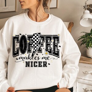 May include: White sweatshirt with the text "COFFEE makes me NICER" in a black, outlined font. The word "COFFEE" has a lightning bolt with a checkered pattern. The design includes small daisy and coffee bean graphics.
