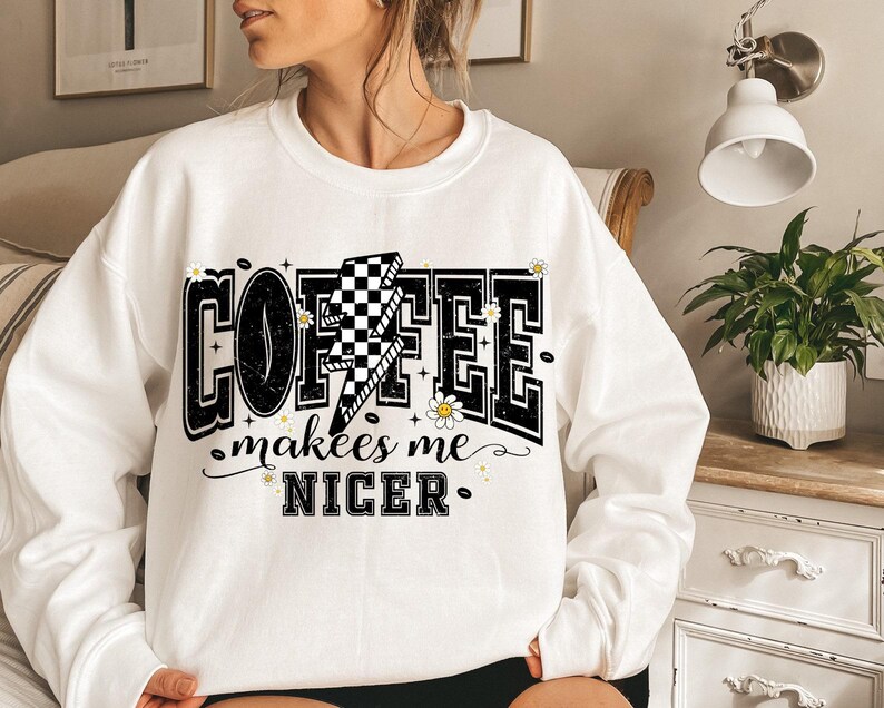 May include: White sweatshirt with the text "COFFEE makes me NICER" in a black, outlined font. The word "COFFEE" has a lightning bolt with a checkered pattern. The design includes small daisy and coffee bean graphics.