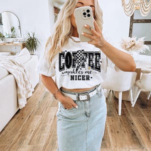 May include: White t-shirt with the text "COFFEE makes me NICER" in a black and white design. The graphic includes a checkered flag and lightning bolt. The person is wearing a light blue denim skirt and a white belt.