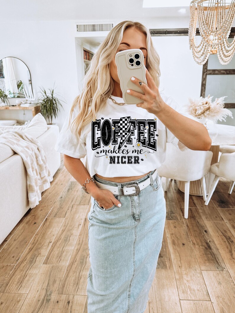 May include: White t-shirt with the text "COFFEE makes me NICER" in a black and white design. The graphic includes a checkered flag and lightning bolt. The person is wearing a light blue denim skirt and a white belt.
