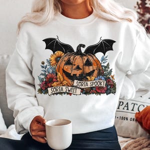 May include: White sweatshirt with a graphic of a jack-o'-lantern with bat wings and flowers. The text "Sorta Sweet" and "Sorta Spooky" is on a banner around the pumpkin.