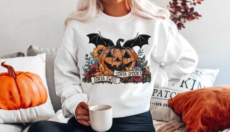 May include: White sweatshirt with a graphic of a jack-o'-lantern with bat wings and flowers. The text "Sorta Sweet" and "Sorta Spooky" is on a banner around the pumpkin.
