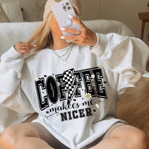 May include: A light gray sweatshirt with the text "COFFEE makes me NICER" in a black, white, and yellow design. The design includes a lightning bolt with a checkered pattern and small daisy accents. The sweatshirt is part of a casual clothing collection.