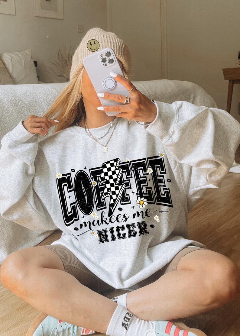 May include: A light gray sweatshirt with the text "COFFEE makes me NICER" in a black, white, and yellow design. The design includes a lightning bolt with a checkered pattern and small daisy accents. The sweatshirt is part of a casual clothing collection.