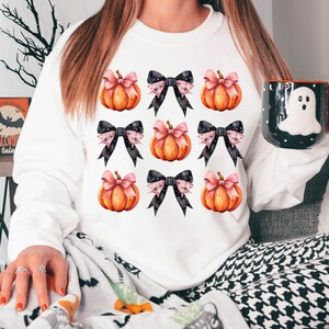 Puede incluir: Sudadera blanca con un patr&oacute;n de calabazas naranjas con lazos rosas y negros.