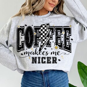 May include: A light gray sweatshirt with the words "COFFEE makes me NICER" in a black, bold font. A lightning bolt with a checkered pattern is in the center of the word "COFFEE". Small daisy graphics are scattered around the text.