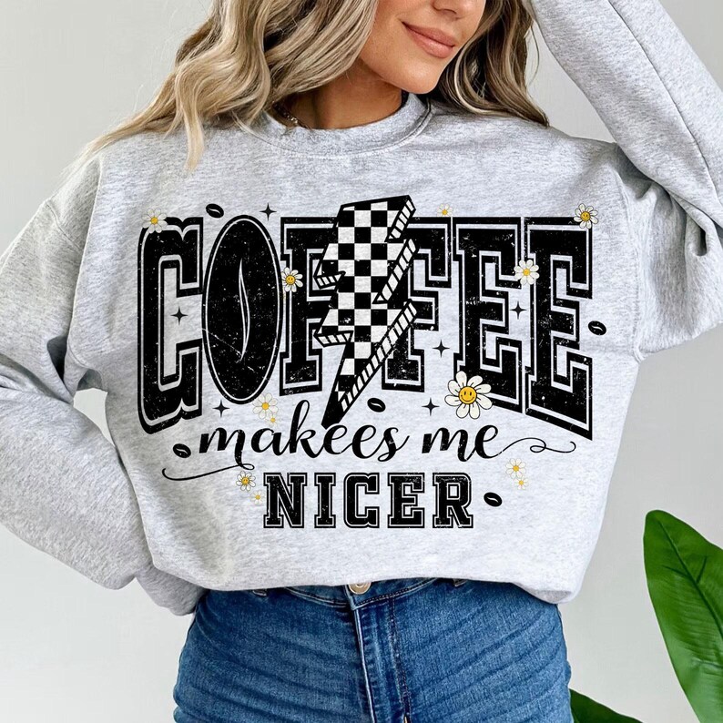 May include: A light gray sweatshirt with the words "COFFEE makes me NICER" in a black, bold font. A lightning bolt with a checkered pattern is in the center of the word "COFFEE". Small daisy graphics are scattered around the text.