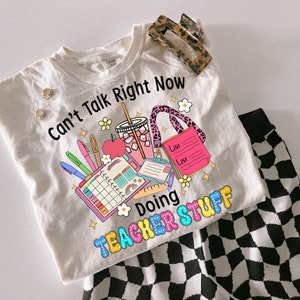 May include: A cream-coloured t-shirt with the text "Can't Talk Right Now Doing Teacher Stuff" and a colourful graphic of school supplies. Includes a leopard print hair clip, stud earrings, and black and white chequered trousers.
