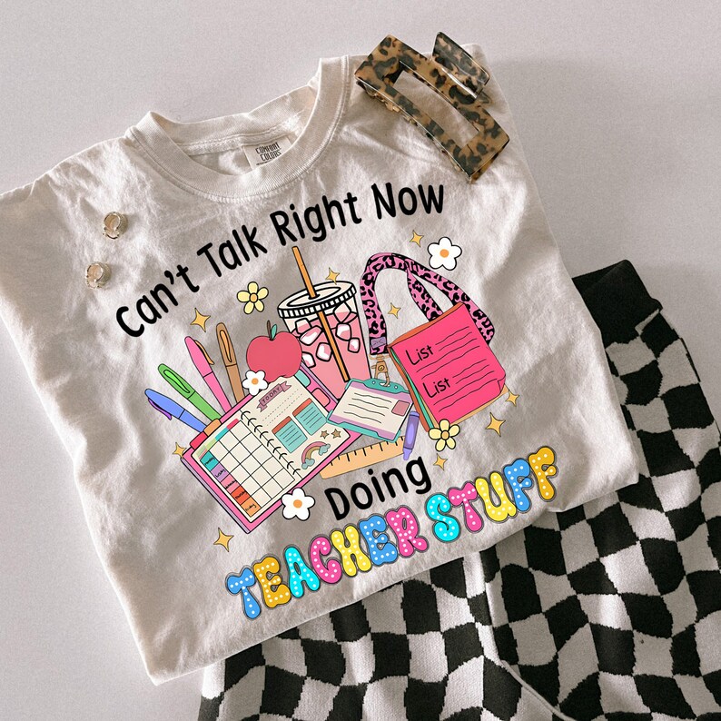 May include: A cream-coloured t-shirt with the text "Can't Talk Right Now Doing Teacher Stuff" and a colourful graphic of school supplies. Includes a leopard print hair clip, stud earrings, and black and white chequered trousers.