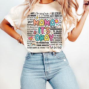 May include: A white t-shirt with a colorful graphic print that says "Mama It's Okay" and includes phrases like "To enjoy the silence", "To set boundaries", "To prioritize your health", and "To not always be strong".